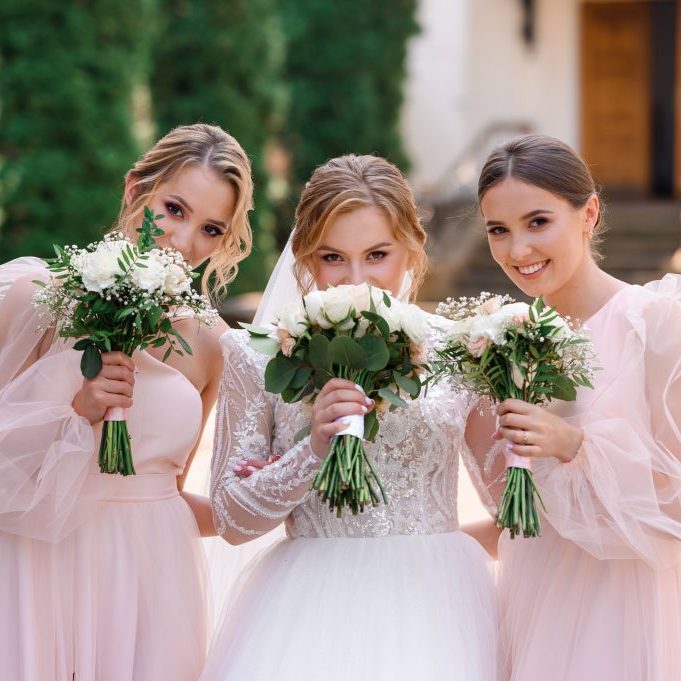 Portrait of elegant bridesmaids and bride in white puffy dress, holding bouquets with flowers waiting for wedding ceremony while posing outdoors.